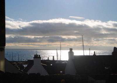 View over roofs towards sunlight reflecting on the sea. Grey clouds lurk beneath blue sky,