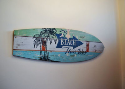 Wooden sign in the shape of a surf board, painted with palm trees, reads: beach this way