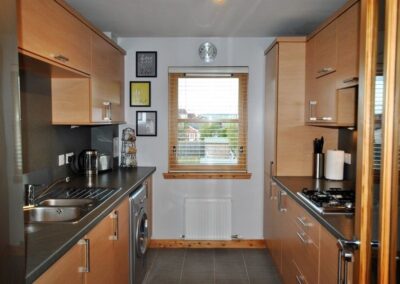 Narrow kitchen with window at far end. Warm, wooden units and silver washing machine