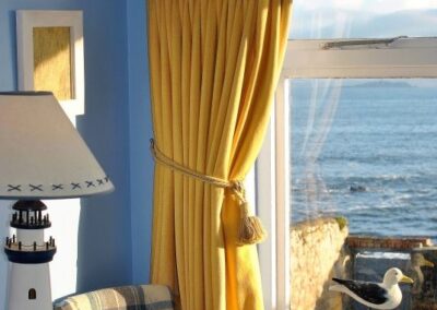 Golden yellow curtain against a blue wall, hanging to the left of a window out to the sea. There is a lamp that looks like a lighthouse on a sideboard.