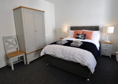 Double bed with wooden bedside tables and lamps. A grey wooden wardrobe sits against a wall next to a wooden chair.