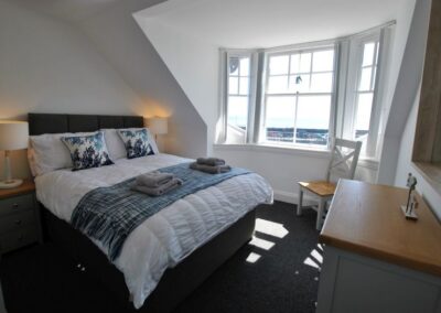 Double bed with wooden bedside tables and lamps. Bay window looks out towards the sea.