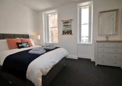 Bed on left, two tall windows and chest of drawers with mirror above