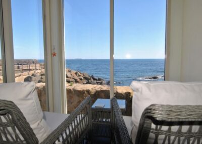 Two wicker chairs with comfortable cushions looking through a large window towards the sea