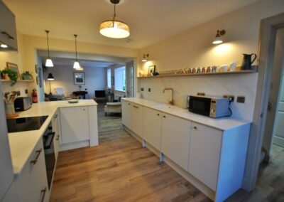 On the left, kitchen counters and worktops with the oven. On the right, counter tops with a sink in the middle. There is a shelf above the sink and downlights.