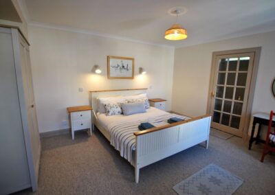 White wooden double bed between bedside tables. Two spotlights on the wall above the bed. To the left of the bed is a door with 15 panes of glass. It is opposite a large wardrobe.