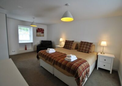 Superking bed with red-brown tartan cushions and rugs in a spacious room. There are windows on either side of the bed.