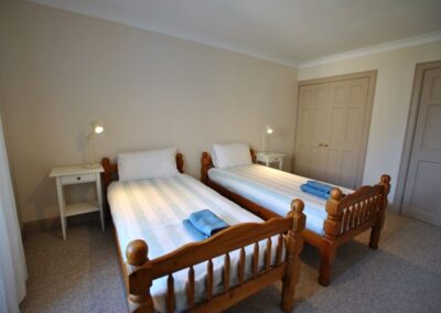 Two wooden beds side by side. There is a bedside table on the left of one and right of the other. There are two sets of doors on the far side of the room.