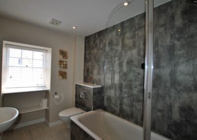 White bathroom suite with shower over bath next to a long, grey feature wall. There is a window opposite the end of the bath.