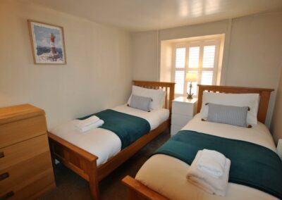 Two single beds with a bedside table between, behind which is a bright window. There is a chest of drawers at the foot of the bed on the left.