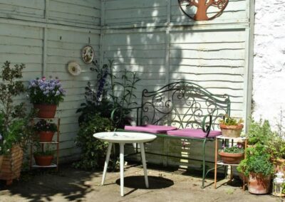 Courtyard corner with light green trellises and potted plants surrounding a thin iron garden bench with purple cushions and a three-legged table.