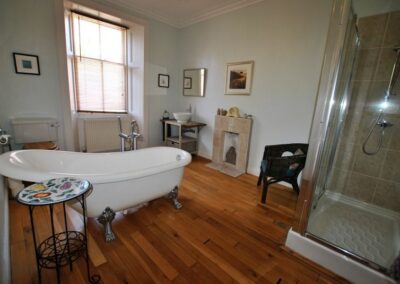 Roll-top bath in centre of wooden-floored bathroom. At the far end is a large window. At this end is a walk-in shower cubicle.