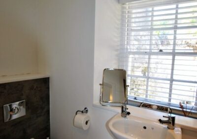 White bathroom suite. There is a window with white venetian blinds opposite the pedestal sink.