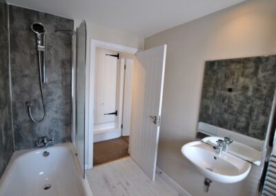 White bathroom suite with grey mottled walls around bath and shower, and above the sink. A wooden door lies open into a hallway with another wooden door with black metal T-hinges up a step opposite.