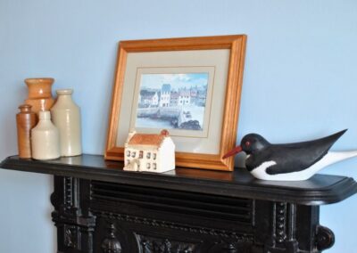 Close-up of objects on a black fireplace. Four earthenware bottles, a painting of a harbour, a small house and a sea bird.