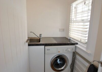 Utility room with window to right of sink. Beneath the counter, to the right of the sink, is a washing machine. A clothes airer sits on the floor beneath the window, nexts to a towel ring.