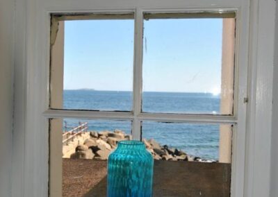 View from a small window out to sea. There is a vase on the windowsill, and balanced on the window is a model of a boat and fishes.