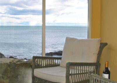 A wicker and cushion chair next to a large window looking out to sea. There is a bottle of wine and two glasses on an occasional table.