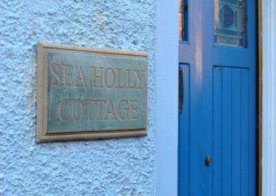 Blue front door with stained-glass inserts. On the wall to the left is a number 5 engraved on a slate, and beneath the house name, Sea Holly Cottage.