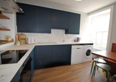 White kitchen with dark units. Dishwasher and washing machine are to the right of the sink, beside a bright window.