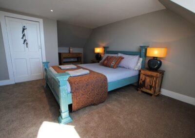 Plenty of space around the turquoise bedframe. There are lamps on bedside tables either side of the bed, and a sloping roof with light coming from an unseen window on the right.