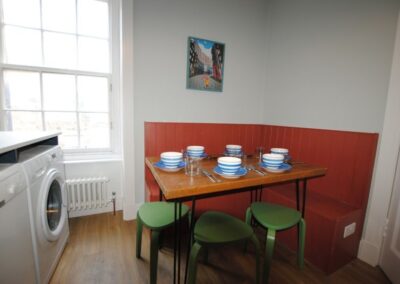 Table set for six in the corner of the kitchen. Three green stools beneath the table plus bench seating against the walls.