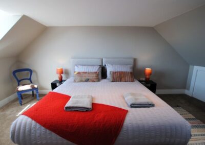 Grey superking bed with red throw and grey towels beneath a sloped roof. Orange bedside lamps either side.