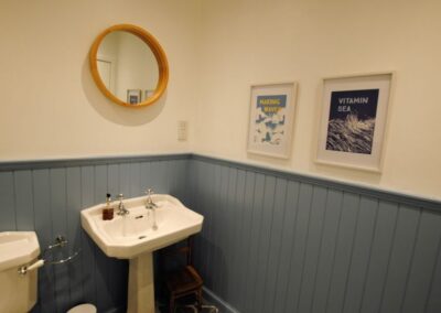 Corner of bathroom with white sink, circular mirror and two framed, sea-themed prints.