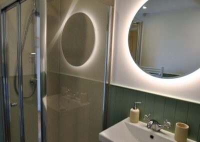 Rectangular walk-in shower in corner of room. To its right is a white sink unit and above that a backlit circular wall-mounted mirror.