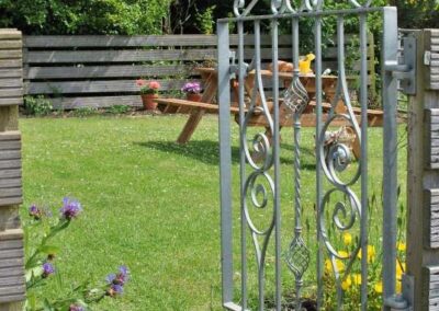 An ornate silver metal gate opens to a garden with a picnic bench in the middle of the grass.