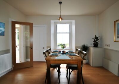 End-on aspect of dining table set for four guests. Beyond the table is a tall window and to the left of that a glass door with wooden frame.