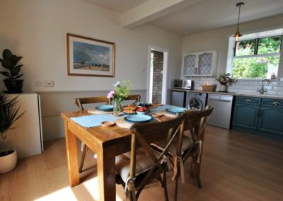 Wooden table set for four guests. Silver fridge, washing machine and dishwasher in the kitchen beyond.