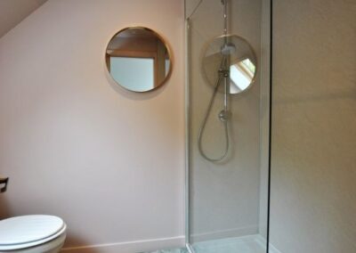 Large walk-in in shower on the right, with a circular mirror on the wall near a white lavatory.