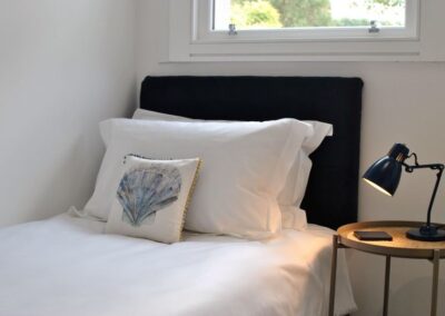Single bed with white linen and a cushion featuring a shell. Behind the headboard there is a window.