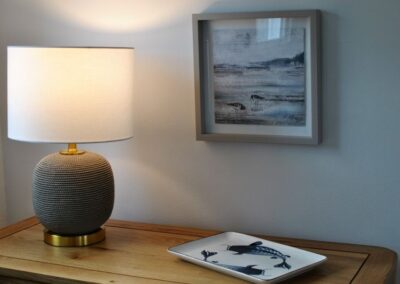Close-up of table lamp next to a plate with a fish design, beneath a seascape painting.