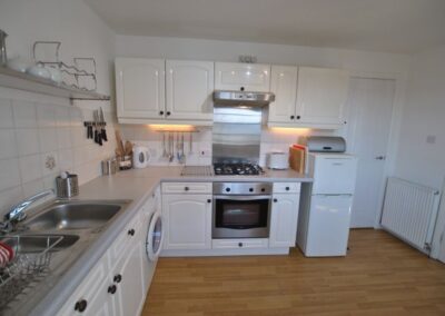 Kitchen with white, glossy cabinets. From left to right: sink, washing machine, hob above oven, fridge freezer with bread bin on top.. There is a door to the right of the fridge.