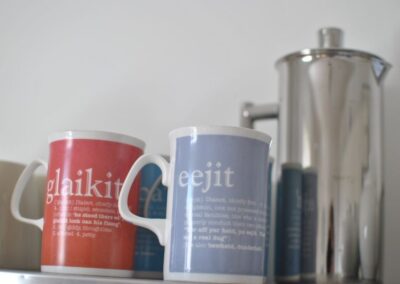 Mugs with Scots phrases: glaikit and eejit with a stainless steel coffee pot.