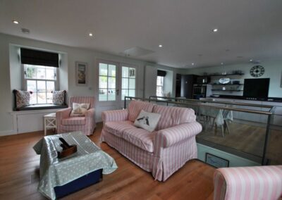 Pink striped sofa and arm chairs. Behind which is stairs down and then a kitchen dining area.