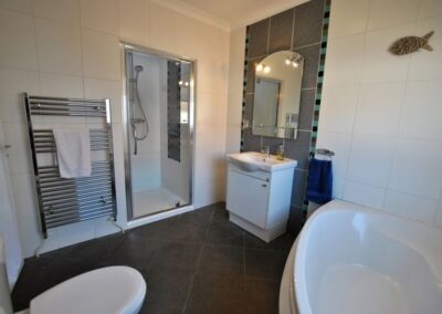 Bathroom with tiled walls and floor. There is a corner bath on the right, and clockwise from there a toilet, heated towel rail, floor-length mirror and sink unit beneath another mirror.