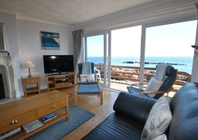 Patio doors open to the sea beyond. Within the lounge there is a large television in the corner to the right of the fireplace.