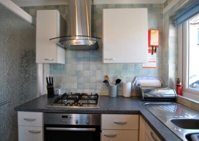 Close up of kitchen with gas hob and extractor unit above, then to the right of it a sink in front of a large window.