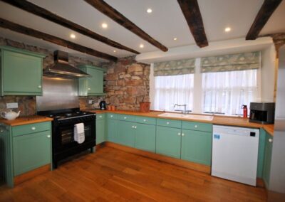L-shaped Kitchen with jade doors. A large gas hob beneath a stainless steel extractor hood. The walls are bare stone, the floor is wooden and the ceiling has beams.
