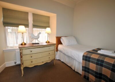 Single bed has an orange tartan blanket. To the left is a chest of drawers with table lamps and a cockerel ornament before a window