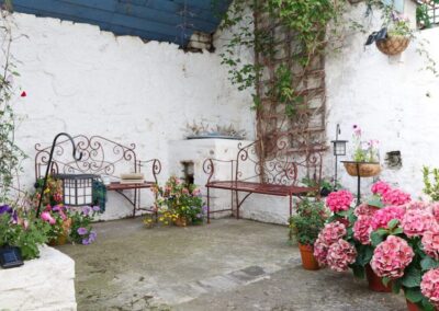 White painted walls decorated with a trellis and hanging basket of flowers. Two ornate benches and plenty of potted flowers.