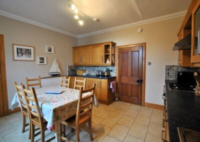 View across dining table towards door and kettle and toaster sitting atop a kitchen counter.