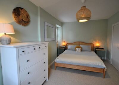 Super kingsize bed in light green room. There is a white chest of drawers on the left with a lamp on top.