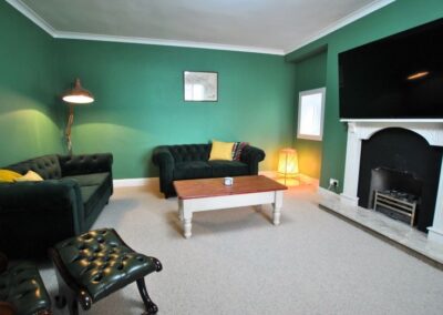 Bright green room with dark green sofas and leather chair and stool. There is a huge TV above a white painted fireplace.