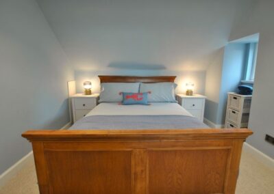 Large wooden framed bed beneath a ceiling that slopes towards the headboard. Either side of the bed is a white bedside table and lamp. There is a small window to the right of the image.