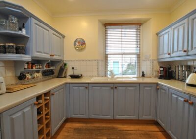 Grey-blue painted kitchen cupboard doors with a magnolia-like countertop and matching walls. There is a window with Venetian blinds in front of the sink.