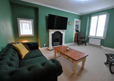 There are two windows on adjacent walls with the fireplace and an alcove in between. There is a long, low coffee table in front oa green sofa.